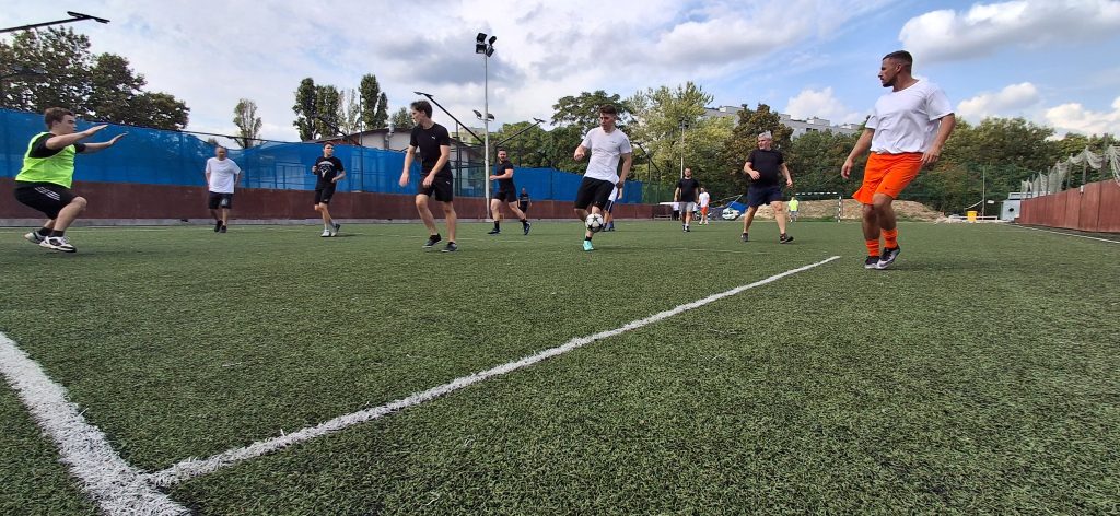 Kispályás futballt játszó férfiak, a kép bal szélén a kapu, a kapus védeni készül. A fotó  apálya sarkáról készült alulnézetből.