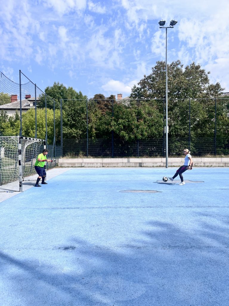 A képen egy nő rűg kapura egy futballabdával. A kép bal szélén a neonsárga pólóban védő férfi kapus, a kép jobb szélén a fehér felsős nő látható. A kép felső felét a fátyolfehős ég foglalja el, süt a nap. 