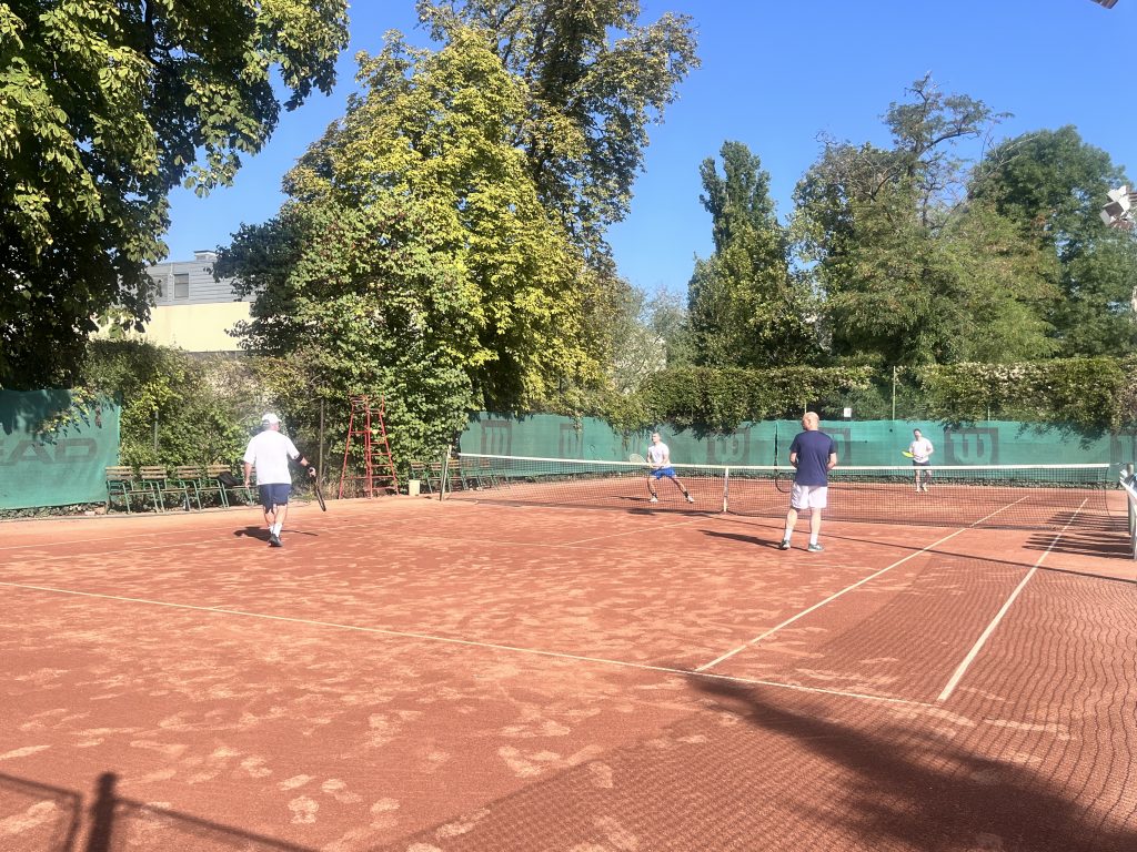 Páros teniszmérkőzés egy napsütötte pályán, négy férfi teniszezik. A háttérben zöldellő fák szegélyezik a salakos teniszpályát.