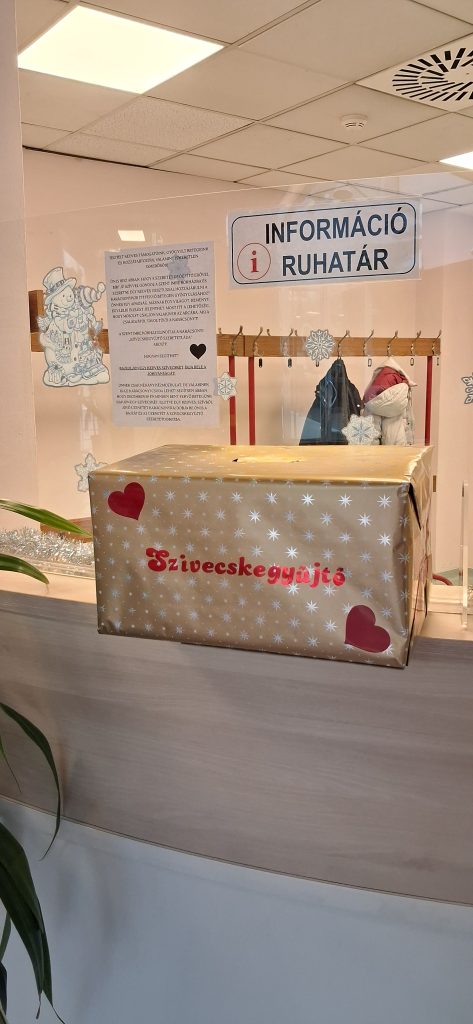 Szívecskegyűjtő Szeretetláda a Szent Imre Kórház aulájában. Arany színű doboz, rajta hópelyhek, és pirossal a Szívecskegyűjtő felirat.